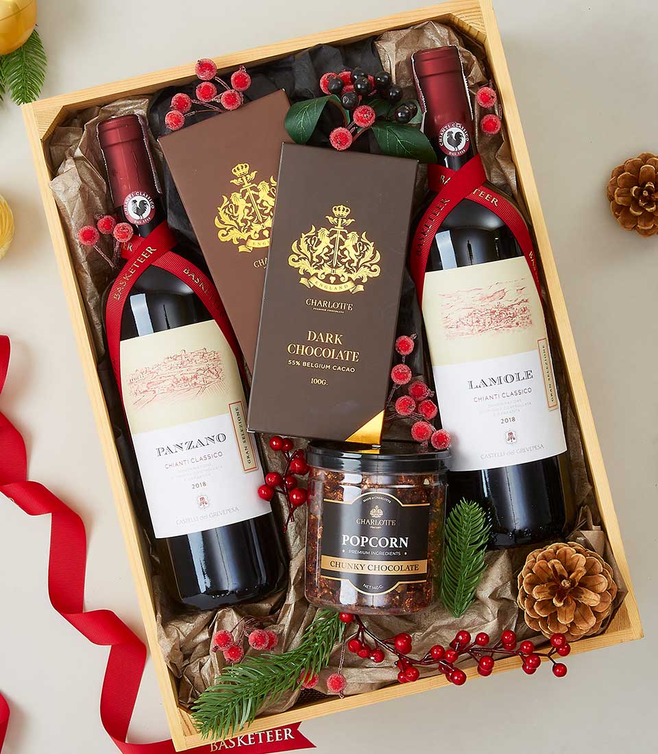 Wooden gift crate with two red wine bottles, chocolate bars, and popcorn nestled in holiday greenery and berries