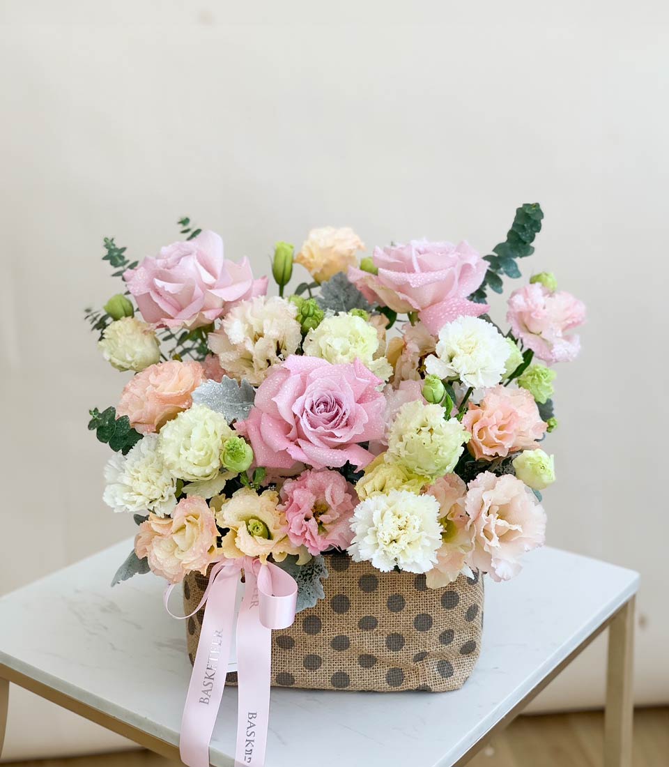 Blooming Bright pastel flower basket with pink roses, carnations, and peach blooms in rustic burlap container