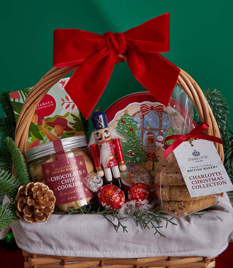 Woven wicker Christmas gift basket with chocolate chip cookies, festive red tin, and British bakery treats tied with red