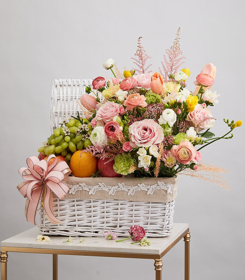 Bright mixed flowers and fresh fruit in white wicker basket with pink ribbon