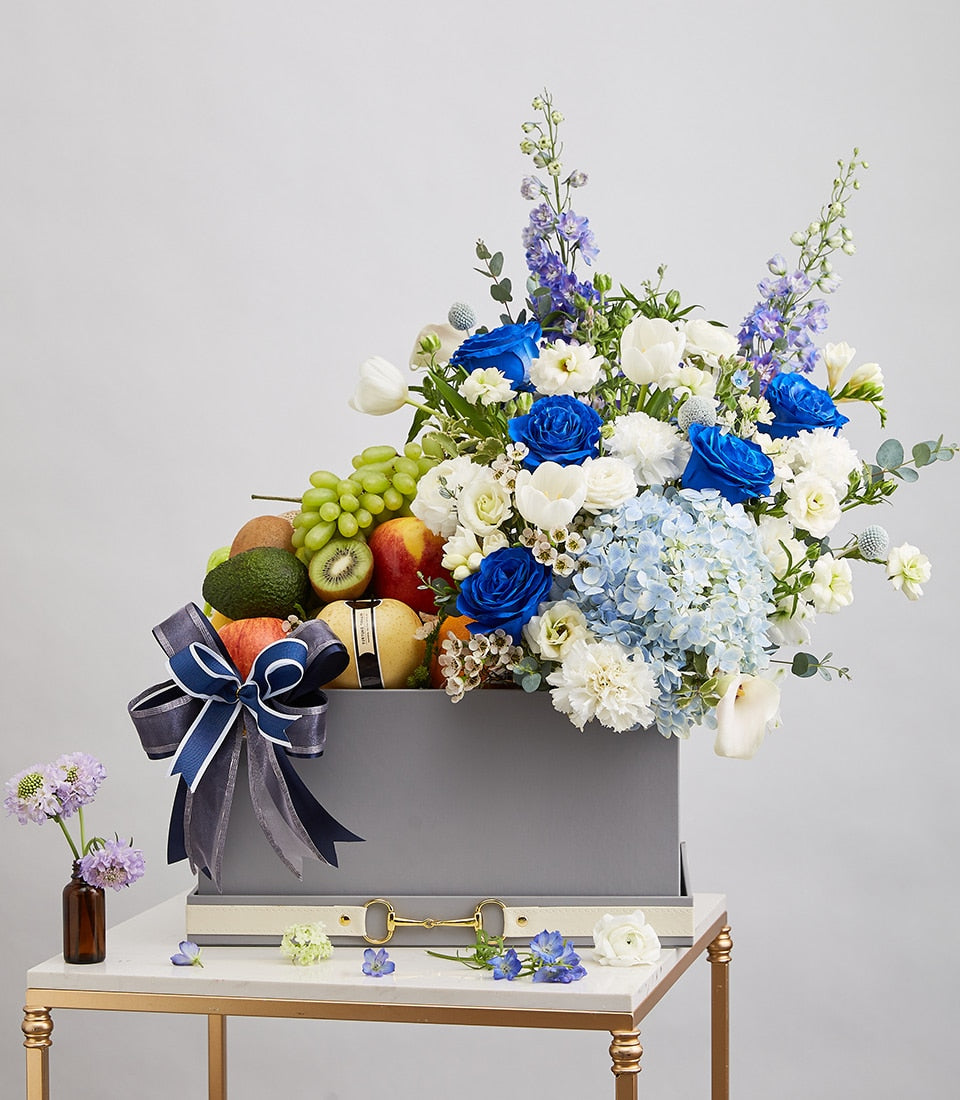Luxury gray gift box with fresh fruit and blue rose bouquet, white tulips, hydrangeas, tied with dark blue ribbon
