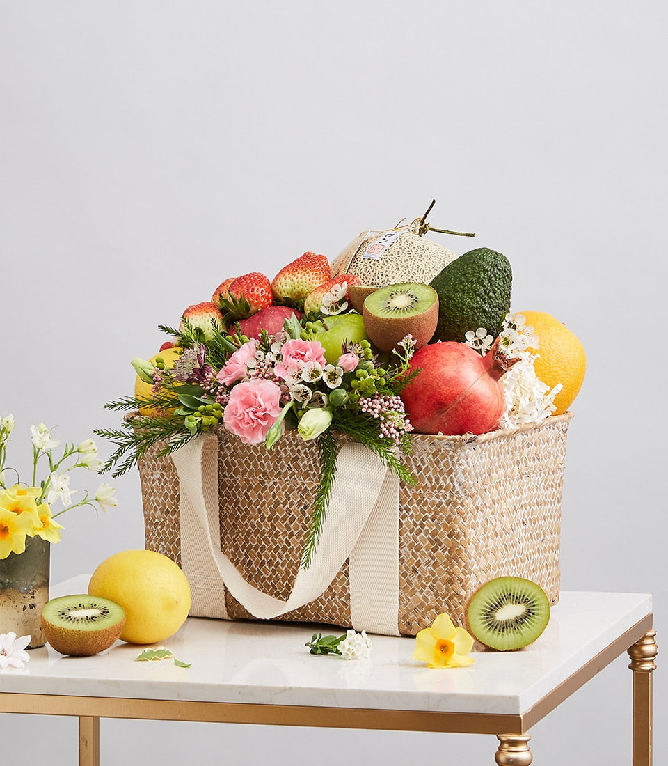 Woven straw gift basket filled with fresh fruits, strawberries, kiwis, apples, and pink carnation flowers with cream handles