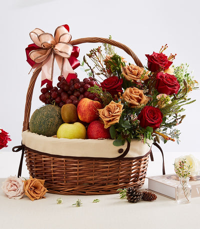 Wicker gift basket with fresh fruits, red and peach roses, and layered pink-red ribbon bow