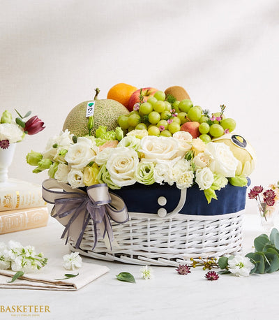 Fresh fruit and rose birthday gift basket with white flowers, green cantaloupe, and orange in woven wicker