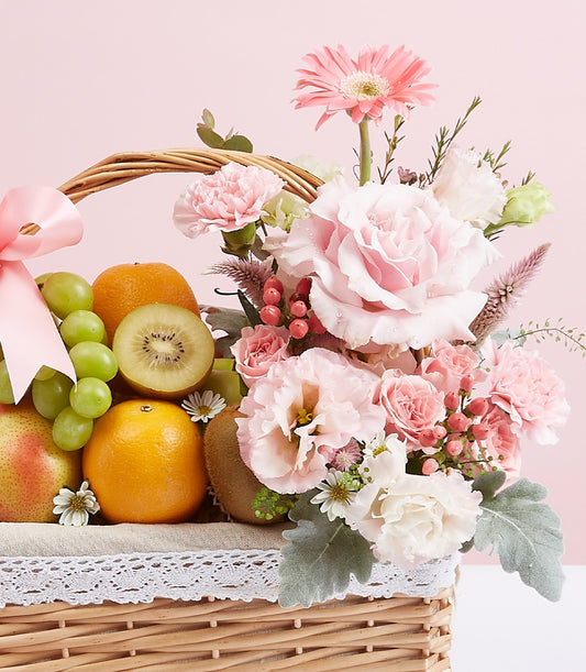 Light brown wicker fruit basket with fresh grapes, oranges, kiwi and pears, topped with pink and white roses and gerbera