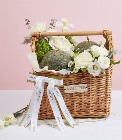 Woven wicker gift basket with fresh green melons, white roses, ranunculus, and premium foliage arrangement