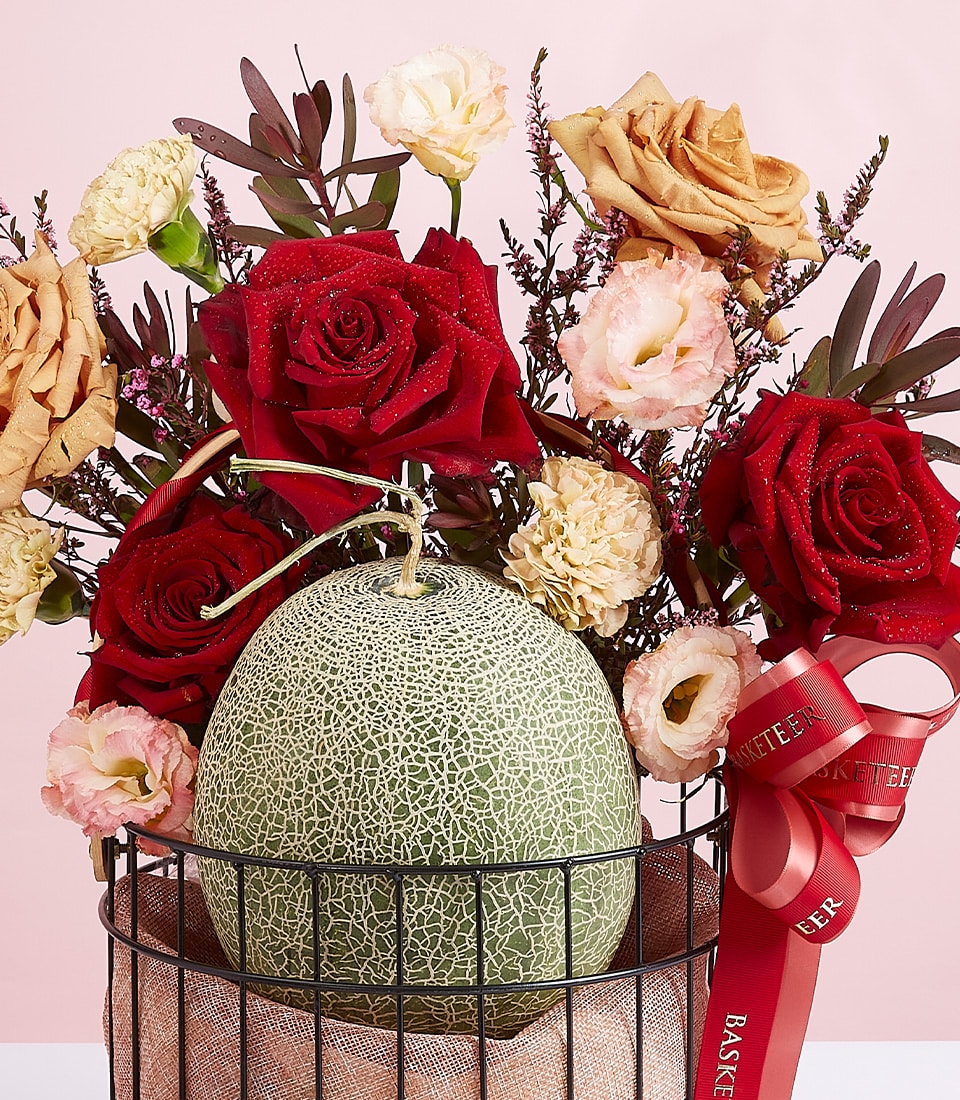 Fresh cantaloupe with vibrant red roses and lisianthus in black wire basket, artisan fruit and flower arrangement