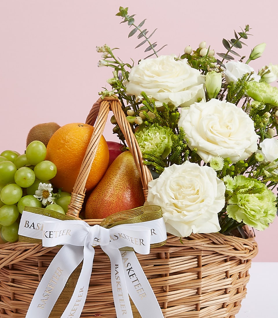 Light brown wicker basket with fresh green grapes, orange, kiwi, and pear arranged with white roses, green carnations, and