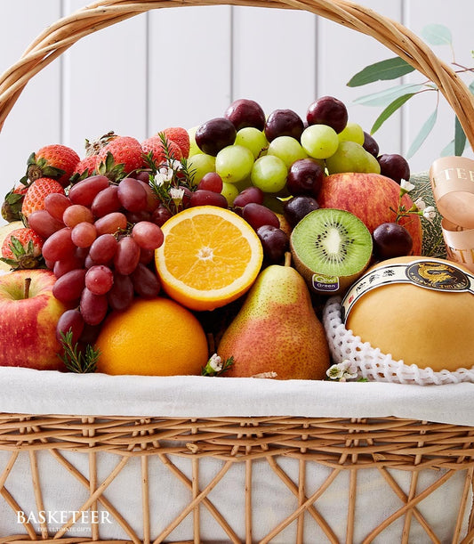 Premium wicker fruit basket with red and green grapes, strawberries, apples, pears, kiwi, orange, and melon, decorated with