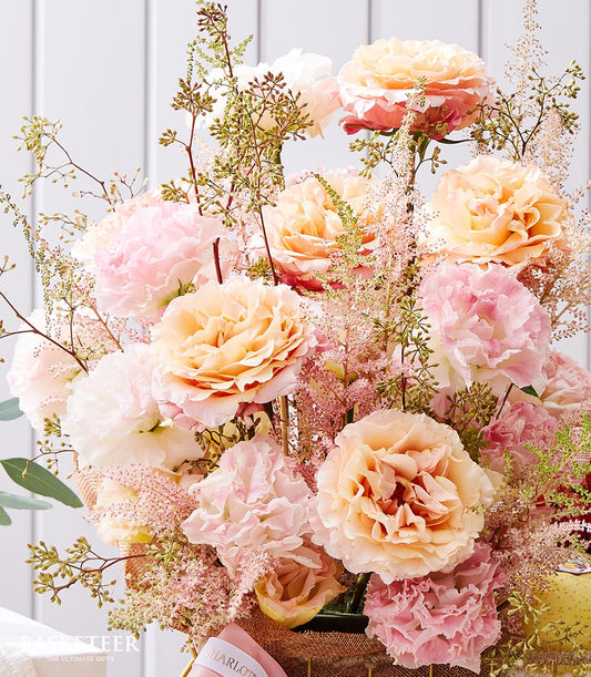 Soft peach and pink garden roses with carnations in burlap-wrapped fruit basket with white ribbon