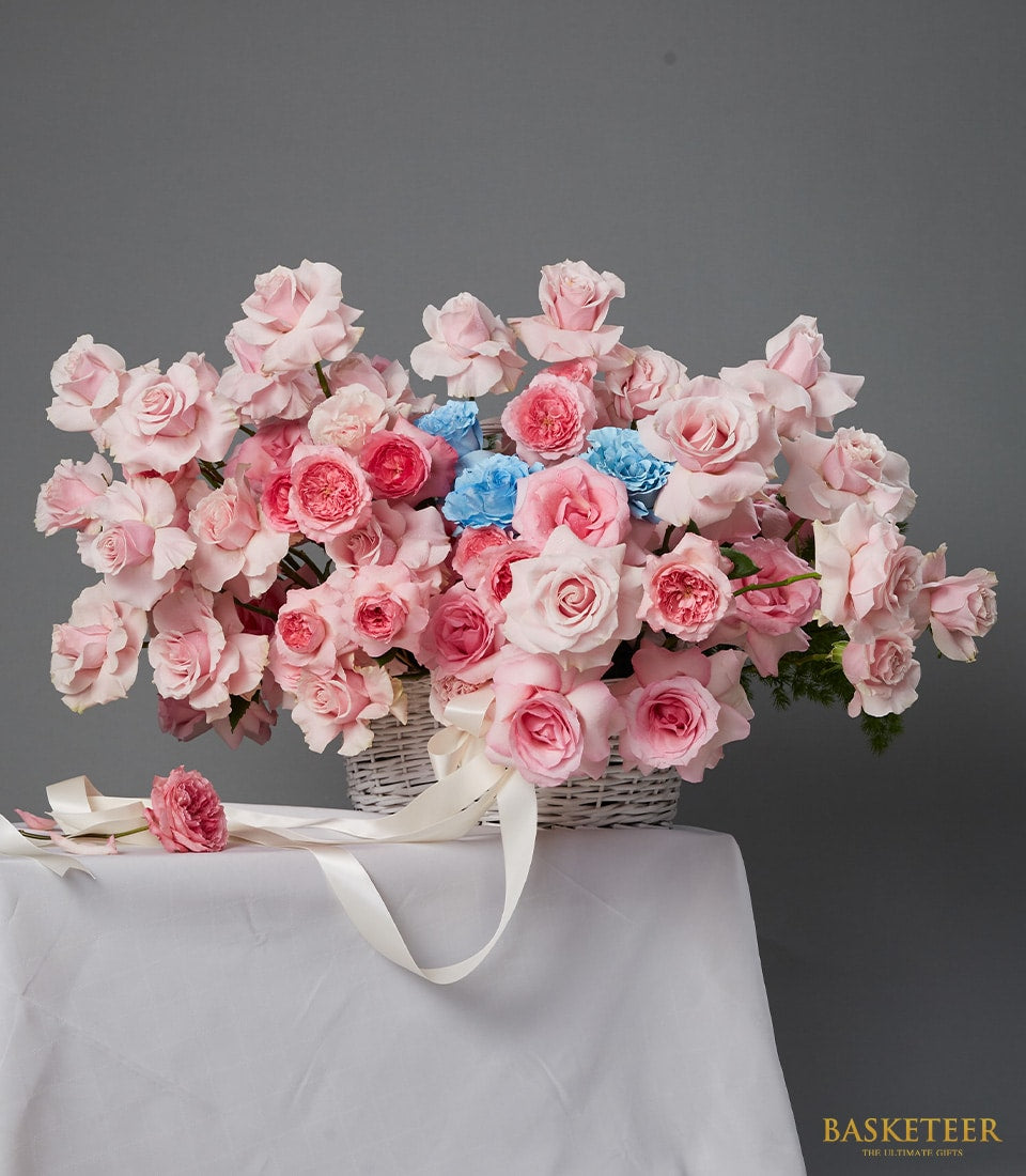 Blush pink roses with blue accents in white woven basket, anniversary flower gift