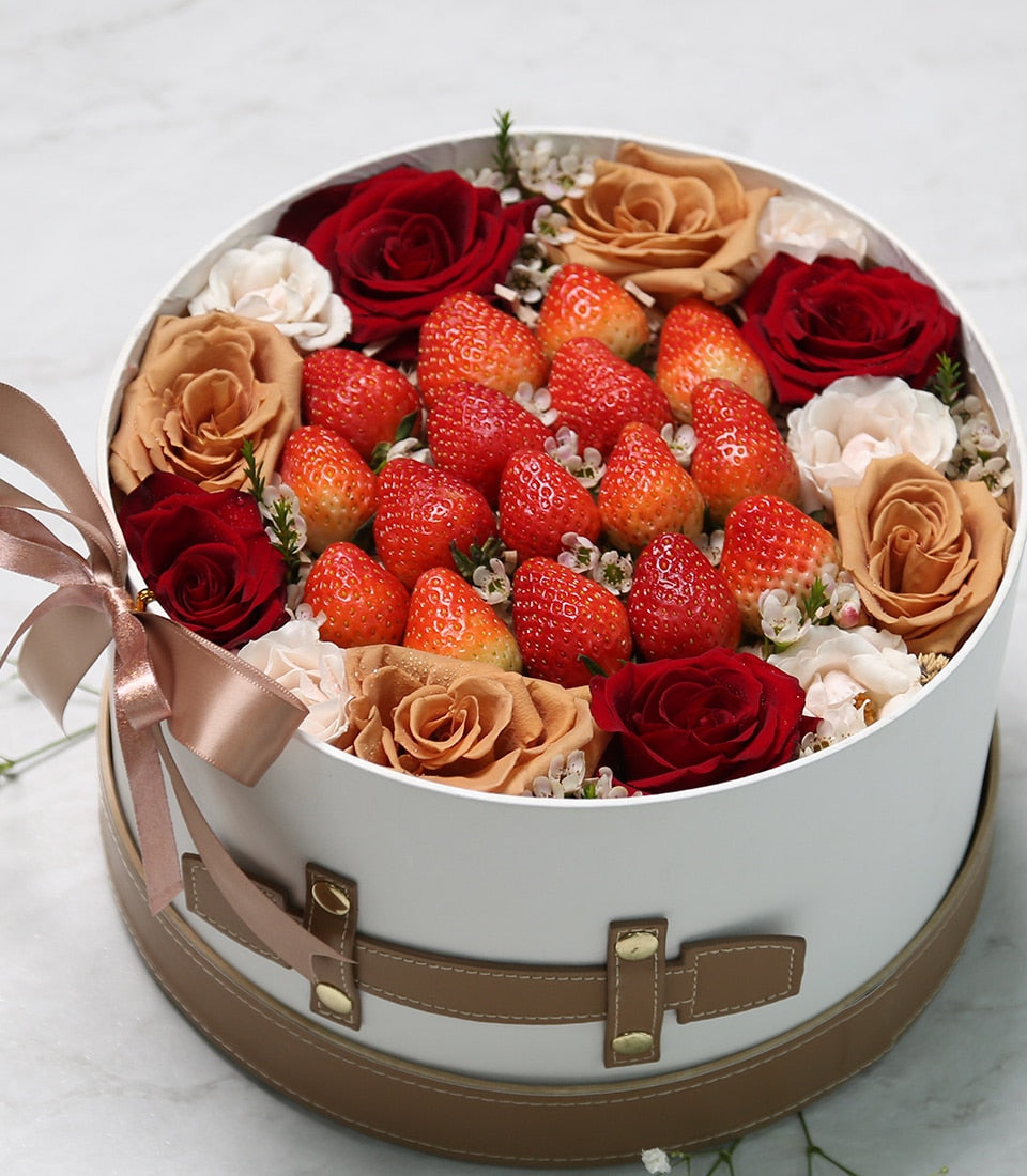 Round white gift box with fresh strawberries, red and peach roses, cream blooms, and faux leather strap with gold studs