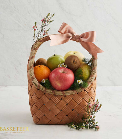 Woven light brown fruit basket with red and green apples, orange, kiwis, pear, peach satin ribbon bow, and white-pink flowers