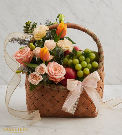 Woven fruit and flower birthday basket with peach roses, orange tulips, grapes, and apples with pink ribbon