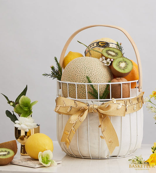 White wire fruit basket with cantaloupe, melon, kiwis, and citrus, tied with gold ribbon bow