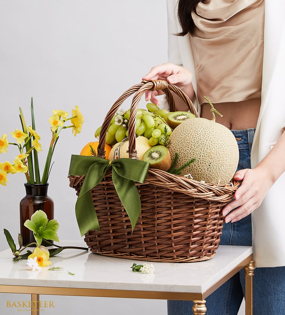 Luxury woven wicker fruit basket with green grapes, cantaloupe, kiwis, pear, and orange tied with dark green satin ribbon