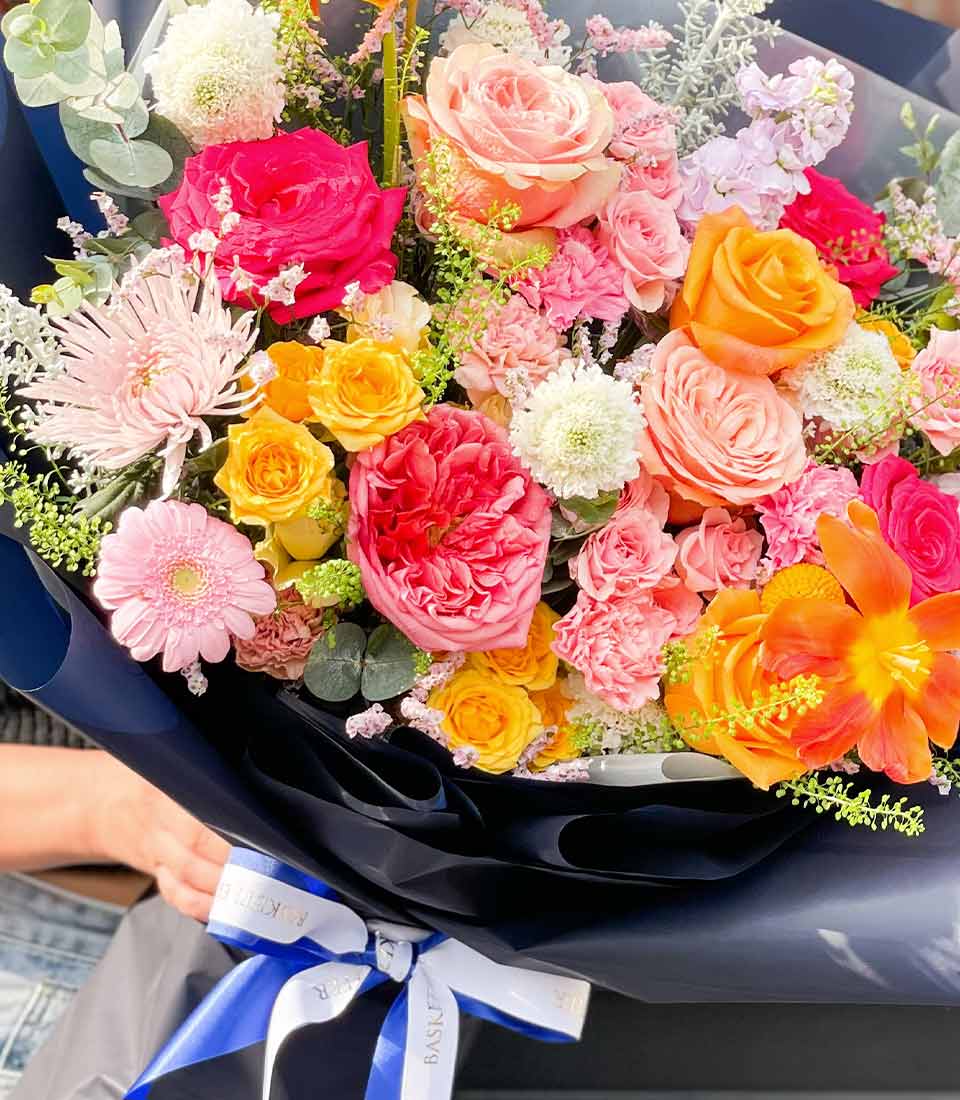 Vibrant mixed rose and daisy bouquet with hot pink, peach, and yellow blooms in navy wrapping