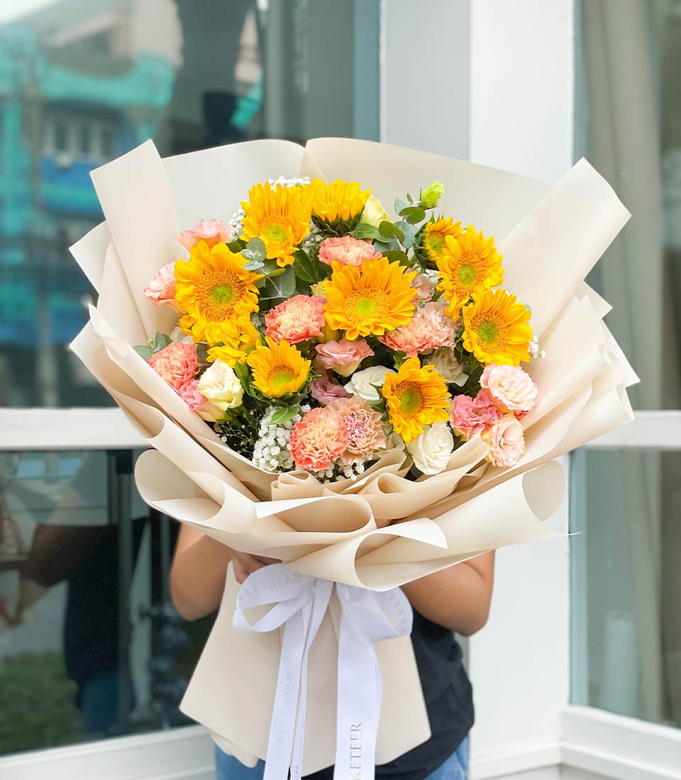 Vibrant yellow sunflower bouquet with pink carnations and white roses, wrapped in beige paper with white ribbon bow