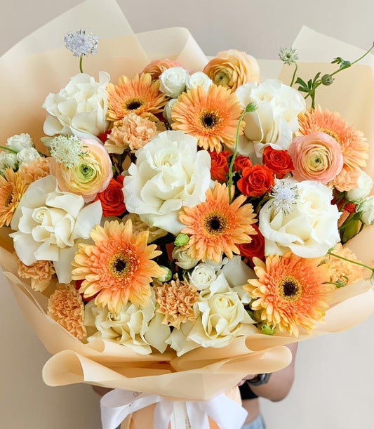 Warm peach and cream rose bouquet with orange gerbera daisies and ranunculus, wrapped in beige paper with white ribbon