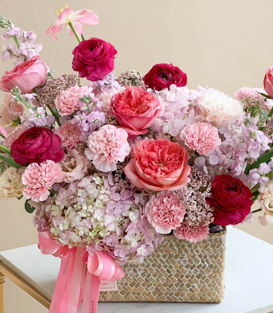 Pink and fuchsia garden roses, ranunculus, and hydrangeas in a woven basket with satin ribbons, congratulations flower gift