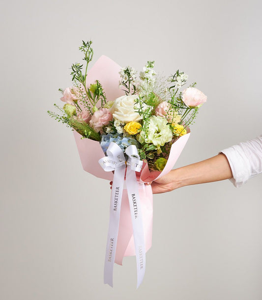 Pastel mixed flower bouquet with white rose, pink carnations, yellow spray roses, and blue hydrangeas in light pink wrapping