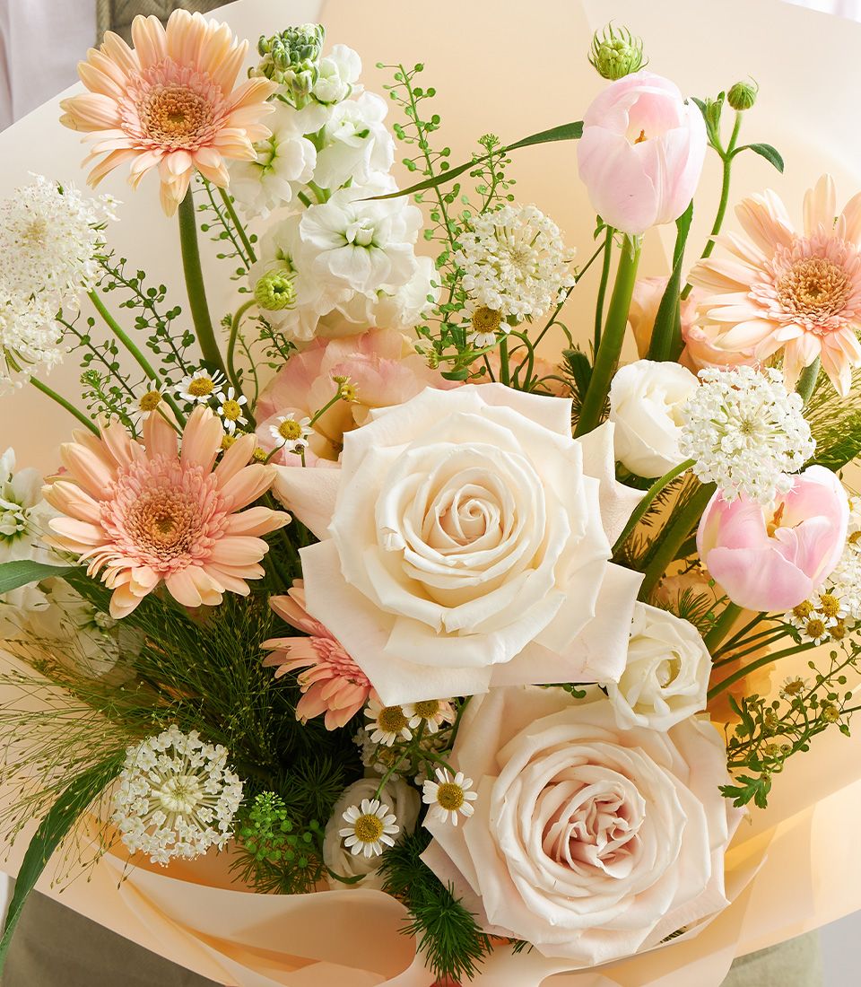Soft pastel mixed bloom bouquet with pink roses, peach gerberas, and white stock flowers wrapped in cream paper
