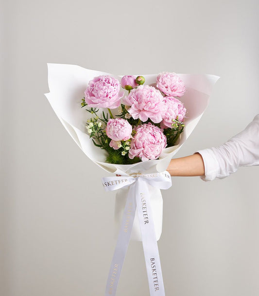 Light pink peony bouquet with white filler flowers and green foliage, wrapped in white paper with gold-ribbon detail