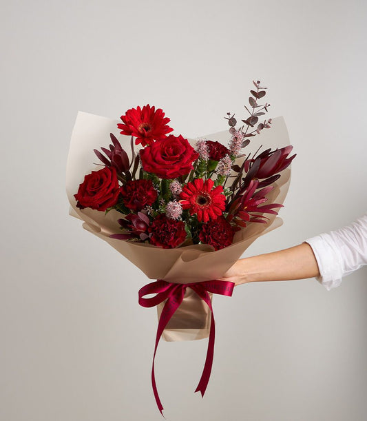 Hand holding passionate red mixed flower bouquet with roses, gerberas, and carnations in kraft paper with red ribbon