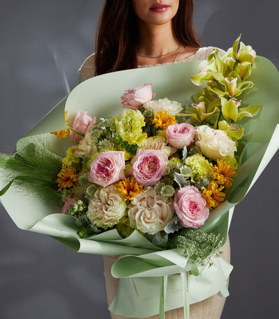 Pastel mixed flower bouquet with soft pink roses, yellow chrysanthemums, and green orchids in mint wrapping