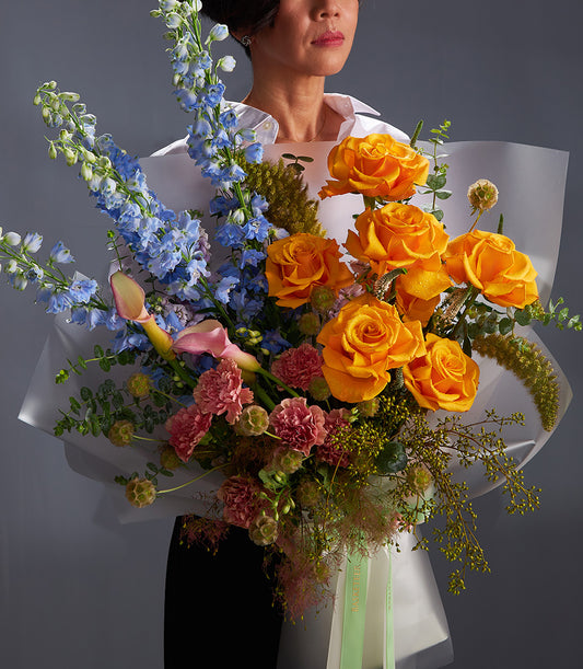 Premium mixed bouquet with orange roses, blue delphiniums, and pink carnations in white wrapping