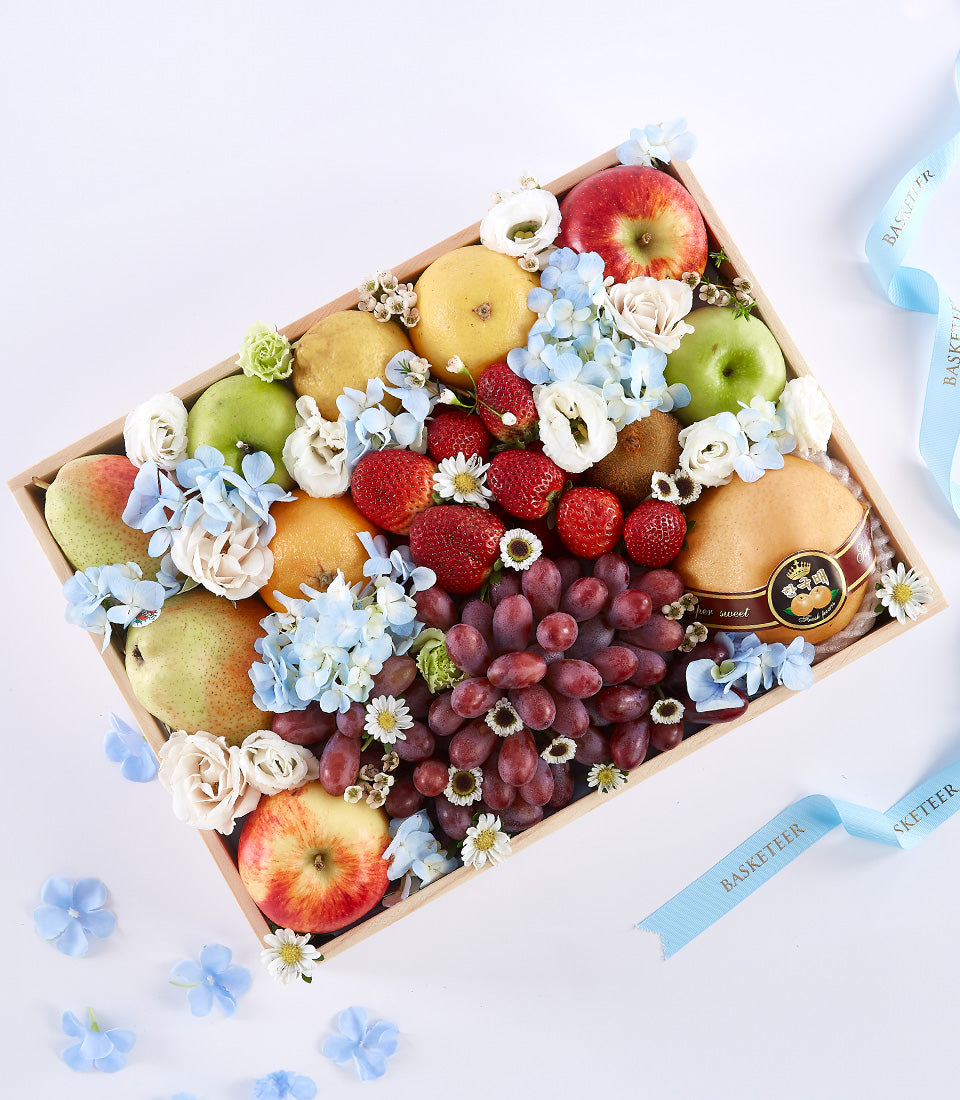 Wooden fruit basket with red and green apples, pears, oranges, grapes, strawberries, kiwi, hydrangeas, roses, and white