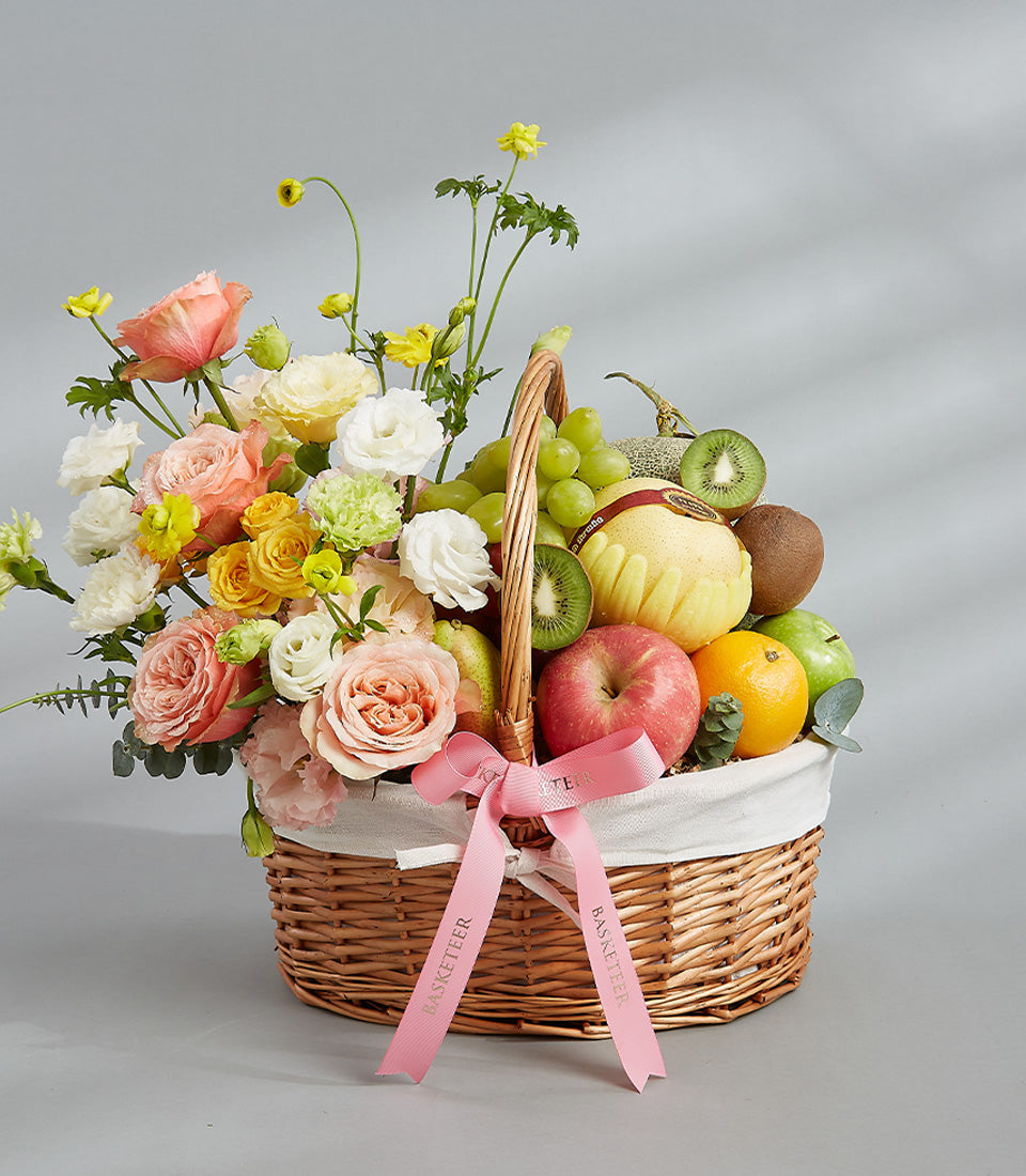 Woven wicker gift basket with fresh mixed fruit, peach roses, ranunculus, and white carnations tied with pink ribbon