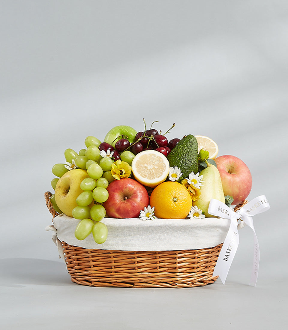 Woven wicker fruit basket with fresh grapes, apples, orange, cherries, and avocado, decorated with white flowers and ribbon