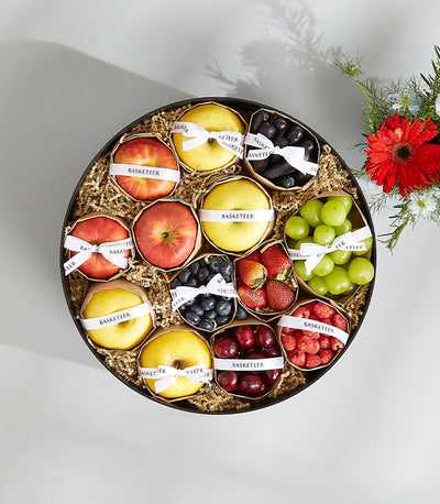 Mixed fresh fruits in circular Basketeer gift box with apples, berries, and red gerbera flowers