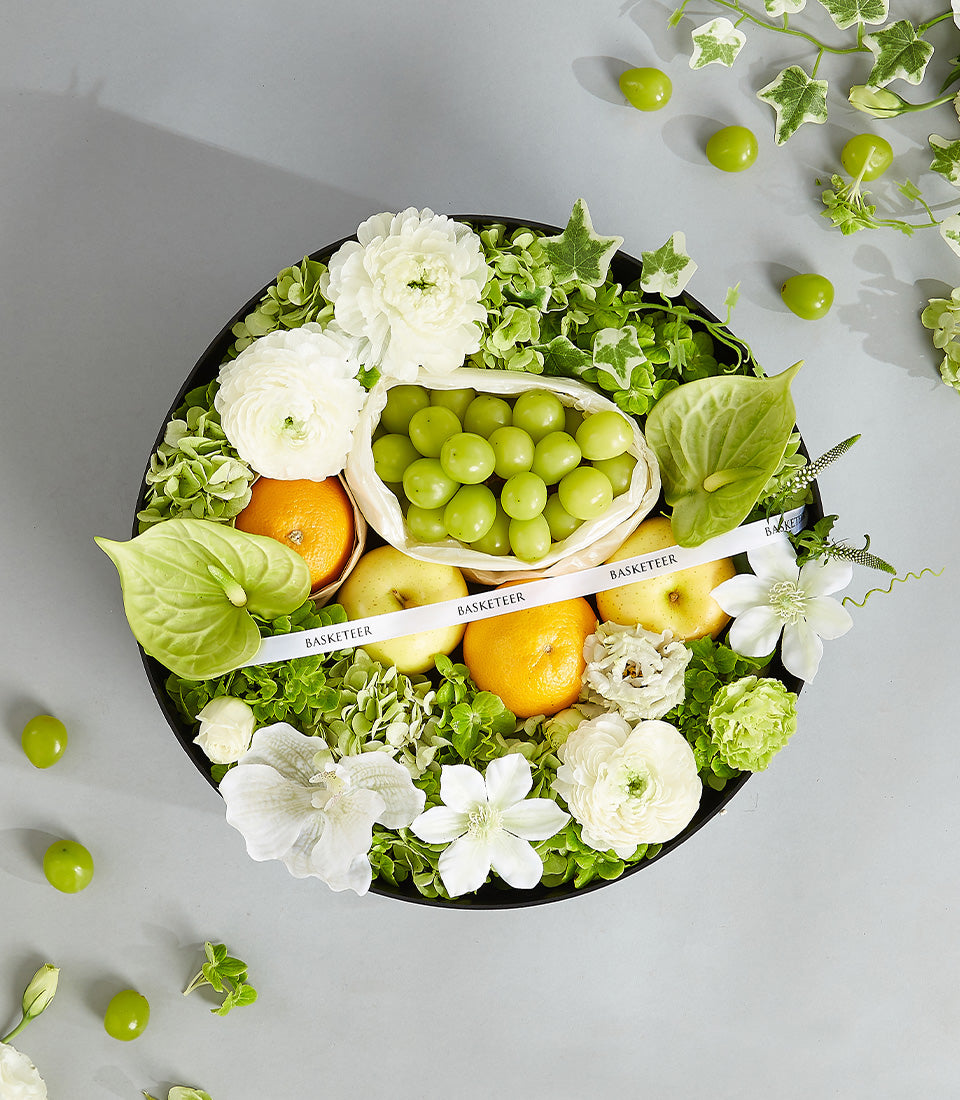 Circular black tray with fresh grapes, oranges, apples, white ranunculus and anthuriums, green foliage, wrapped with