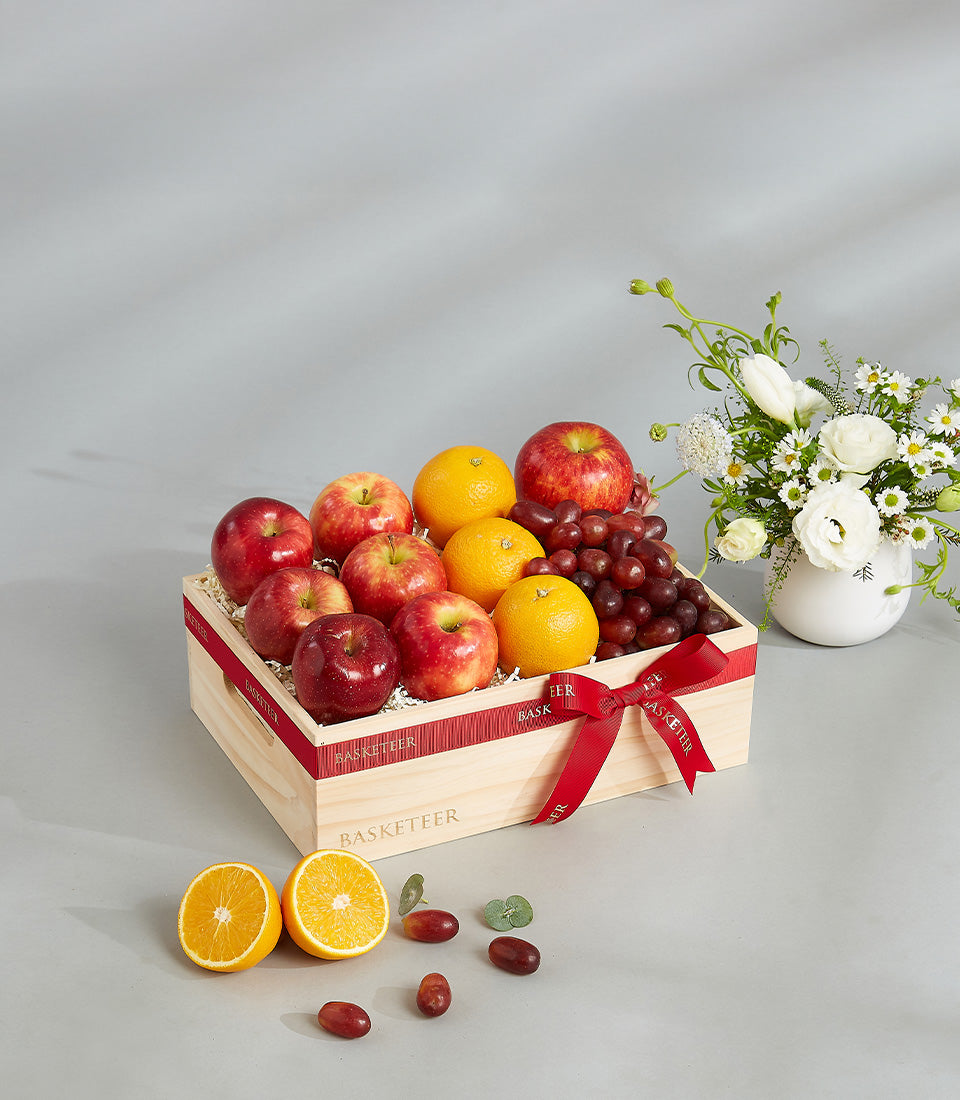 Wooden fruit crate with red apples, oranges, and grapes tied with red ribbon, paired with white tulips and ranunculus