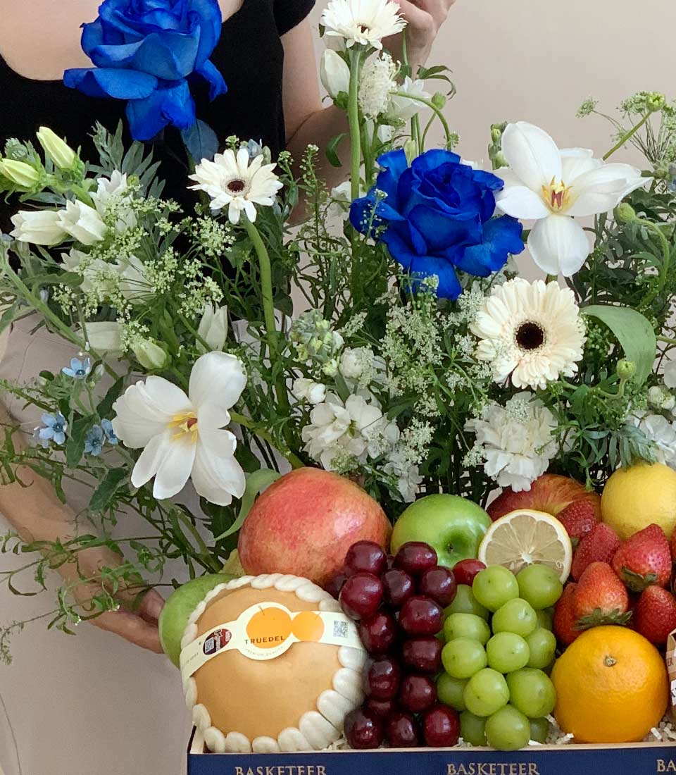 Luxury blue roses and white gerbera daisies with fresh fruit basket featuring pomegranate, apples, and grapes