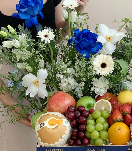 Luxury blue roses and white gerbera daisies with fresh fruit basket featuring pomegranate, apples, and grapes