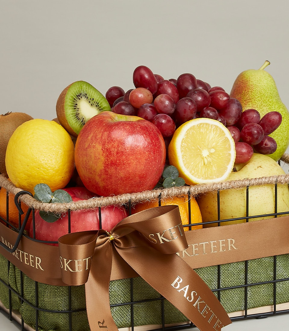 Black wire fruit basket with green burlap lining filled with fresh apples, lemons, kiwis, grapes and pears, tied with satin