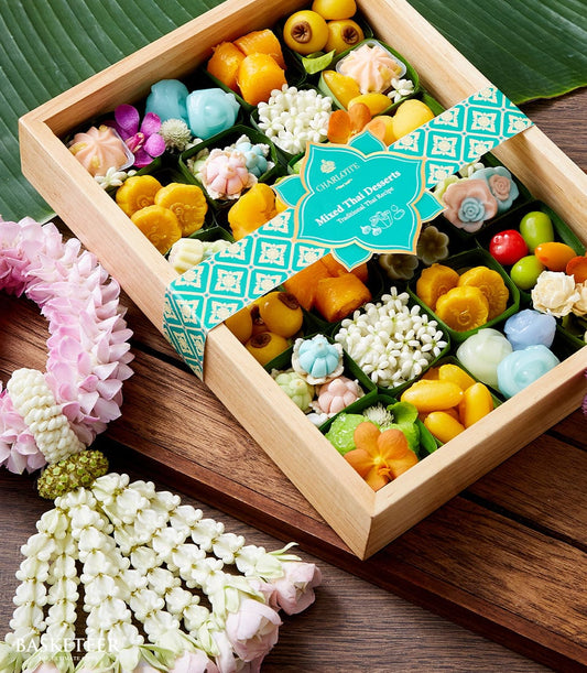 Wooden box of mixed Thai desserts in pastel colors with teal ribbon, paired with white and pink flower garland for Songkran
