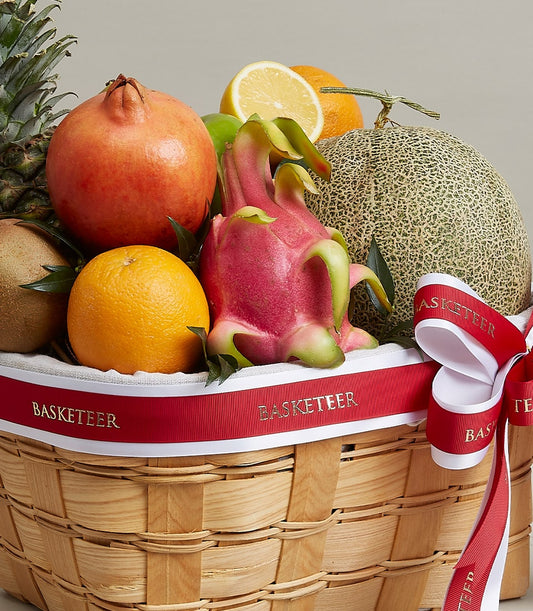 Woven wood fruit basket with pineapple, pomegranate, dragon fruit, and citrus on white fabric lining, red ribbon accent