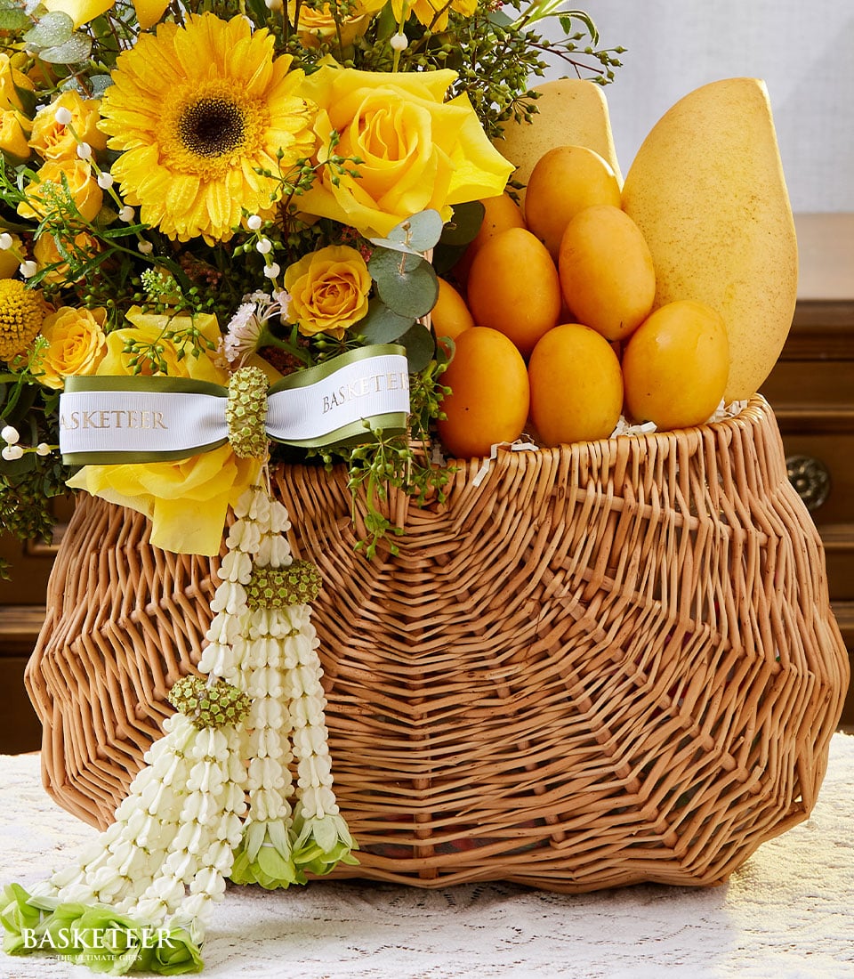 Light brown wicker basket with yellow gerbera daisies, roses, fresh mangoes, and green foliage for Thai New Year gifting