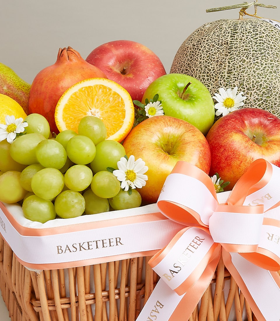 Woven wicker fruit basket with fresh apples, grapes, pomegranate, and melon, tied with white ribbon bow for birthday gifting