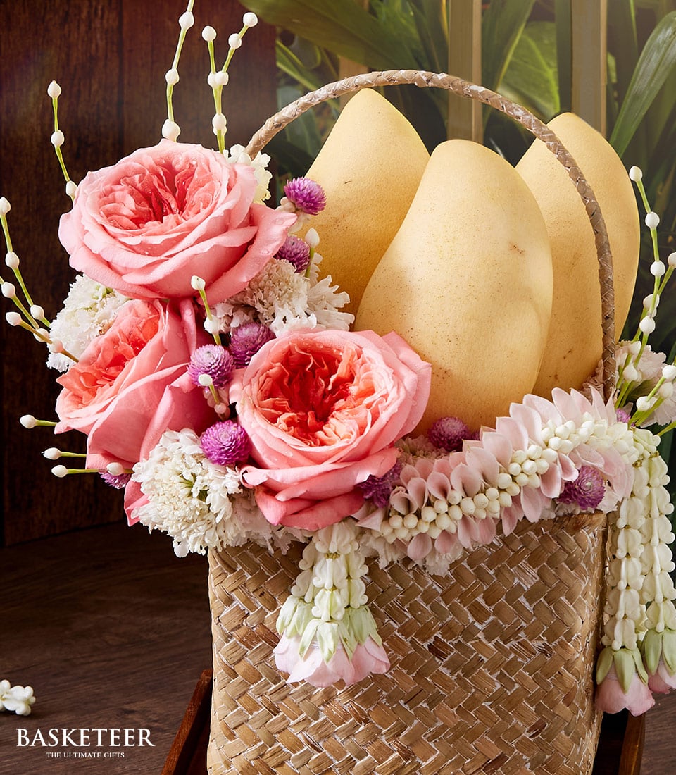 Woven rattan gift basket with fresh pink roses, white flowers, yellow mangoes, and Thai garland for Songkran celebration