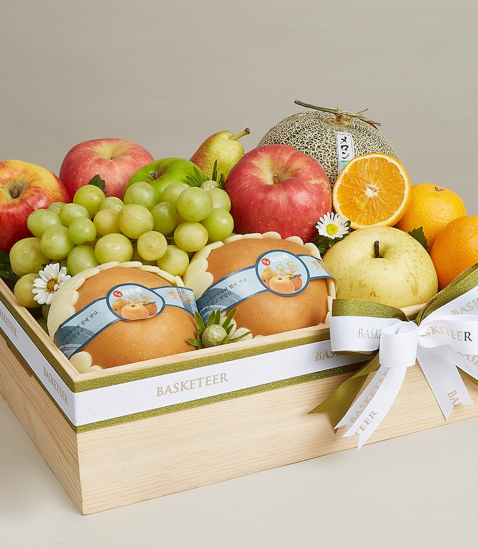 Light wood gift box with red apples, green grapes, pears, oranges and cantaloupe, adorned with white daisies and green ribbon
