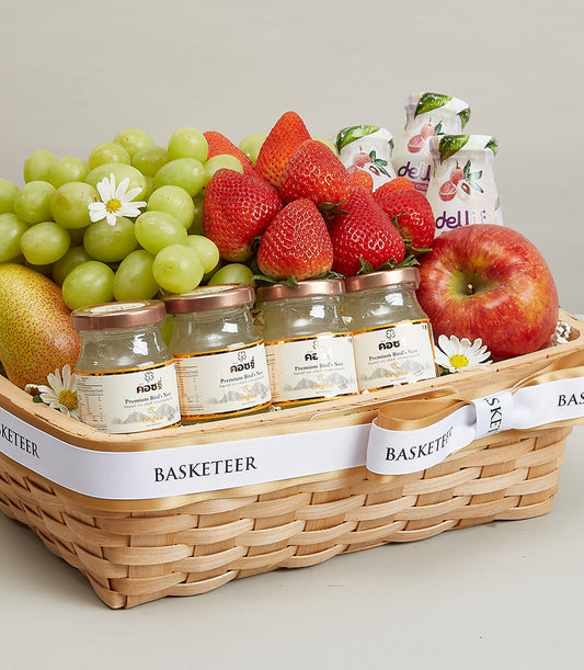 Light brown wicker gift basket with fresh green grapes, strawberries, pear, apple, premium bird's nest bottles, and white