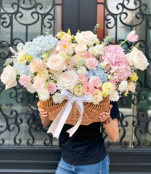 Woven wicker basket overflowing with soft pink roses, hydrangeas, and cream flowers tied with white satin ribbon