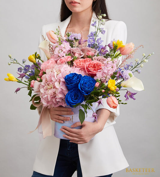 Vibrant mixed flower arrangement in light purple hatbox with pink hydrangeas, blue roses, and garden roses for get well