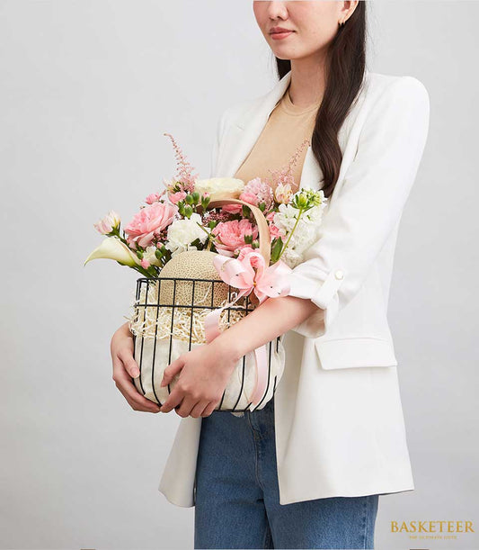 Vibrant melon and fresh flower gift basket with pink roses, white calla lilies, and cream netted melon in black wire frame