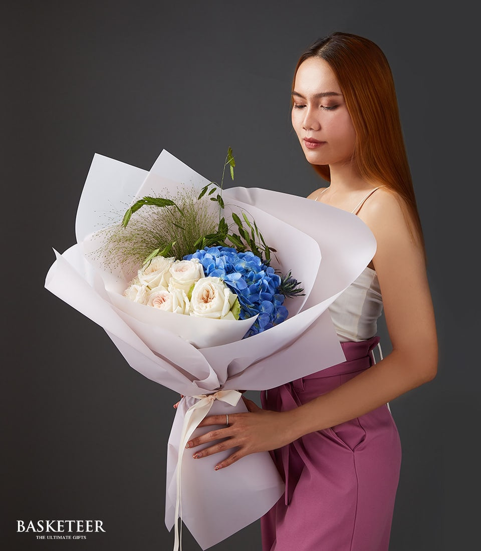 Large bouquet of white and pink roses with vibrant blue hydrangeas, wrapped in white and lavender paper with cream ribbon
